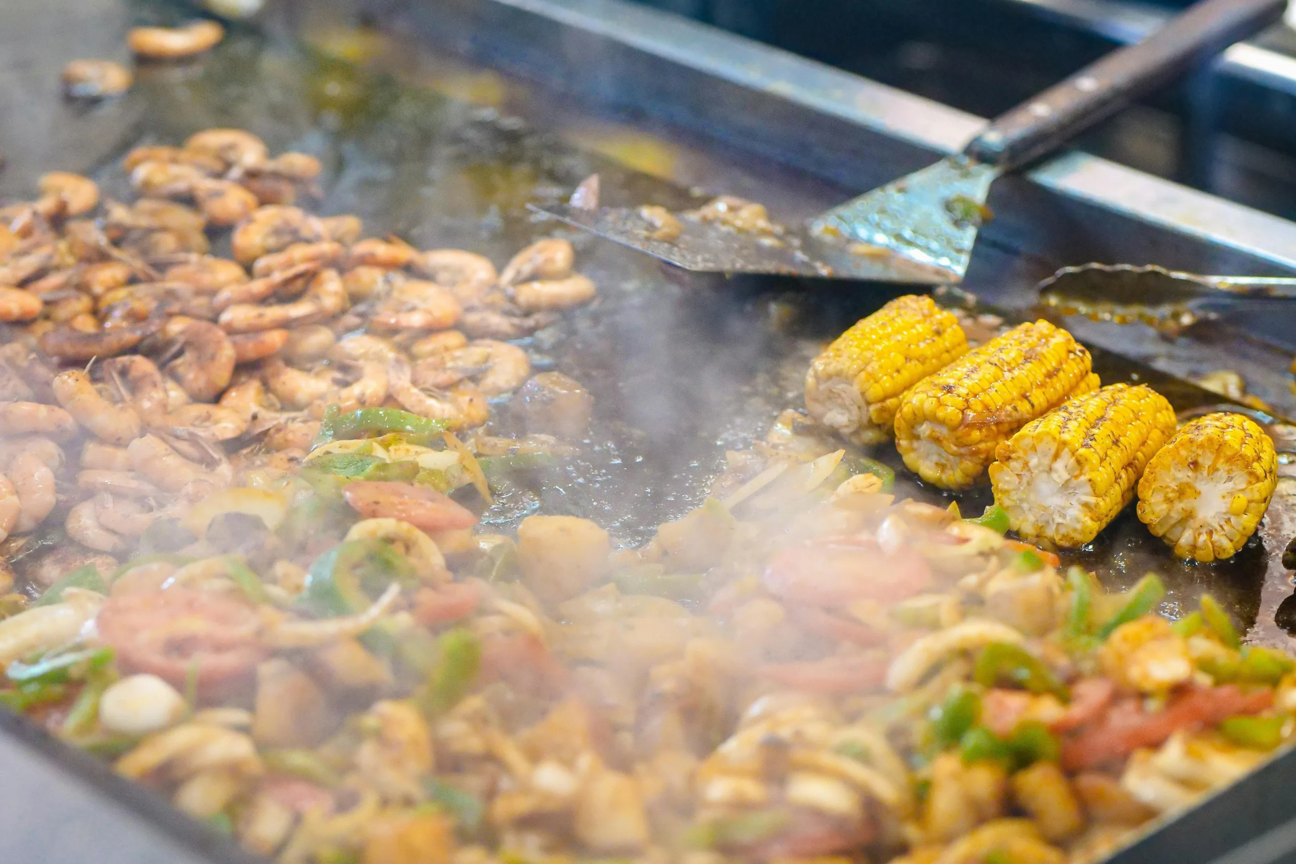 Prawns, peppers, onions, sausage and corn cooking together on a hot BBQ plancha griddle.