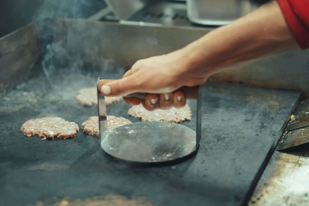 What Is a BBQ Plancha and Is It Worth It for UK Gardens? 2 Hand pressing a smash burger onto a hot BBQ plancha griddle plate.