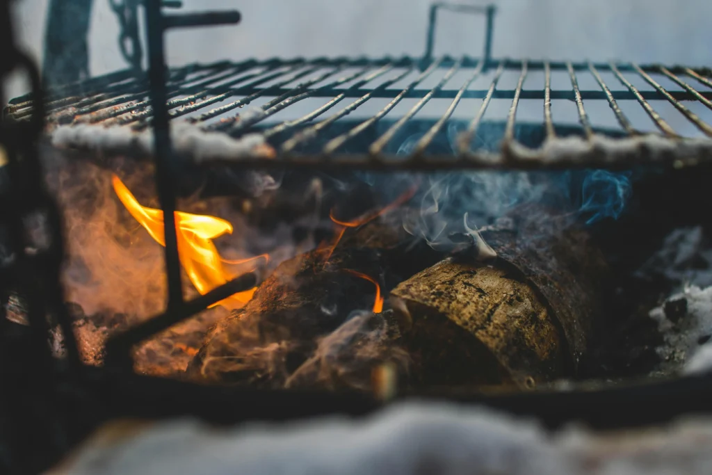 Hardwood logs burning beneath a wood-fired BBQ grate with flame and smoke.