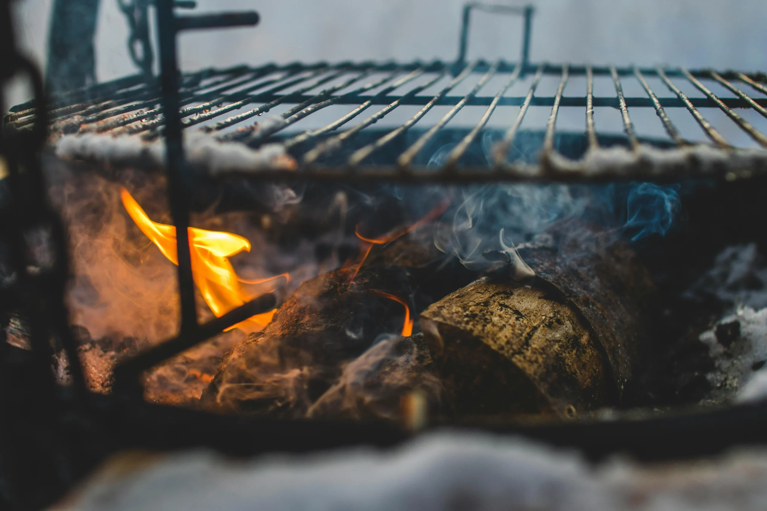 Hardwood logs burning beneath a wood-fired BBQ grate with flame and smoke.